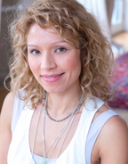 Picture of Lady Samantha, dressed in white with necklaces. She has curly blonde hair on a round heart shaped face. She is of mixed ethnicity. 